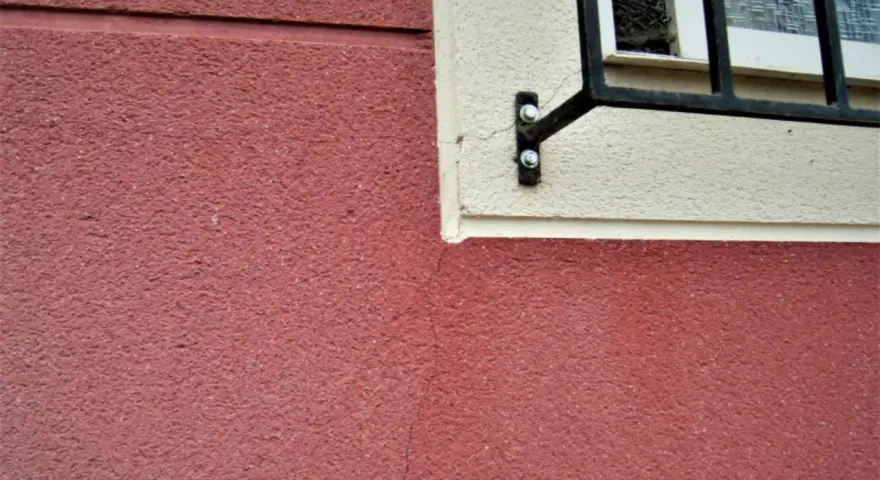 Grietas profundas en pared de color rojo, cerca de una ventana con rejas. Aprende como arreglar grietas profundas en paredes.