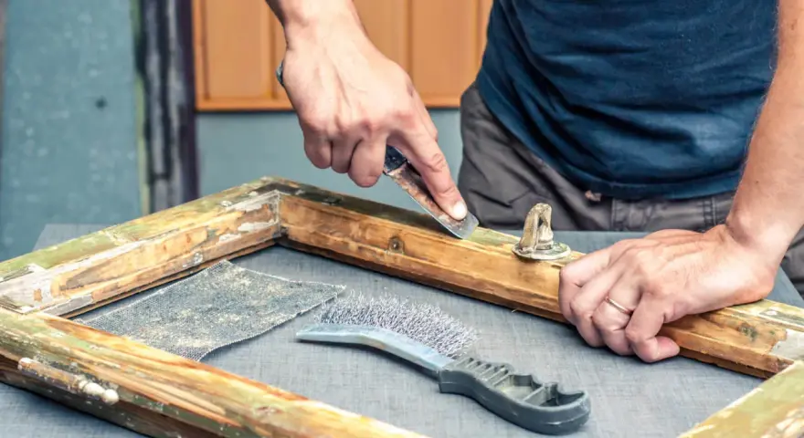 Hombre decapa madera de un marco antiguo con rasqueta y cepillo.