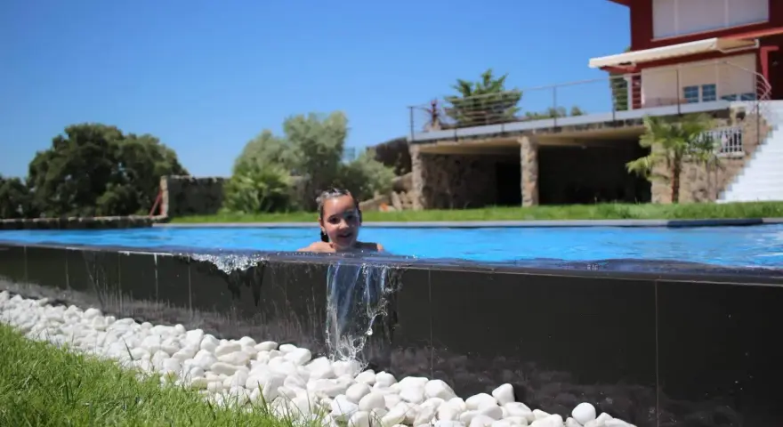Niña sonriente en una piscina desbordante, con agua cayendo sobre piedras blancas y césped verde. Casa moderna al fondo.