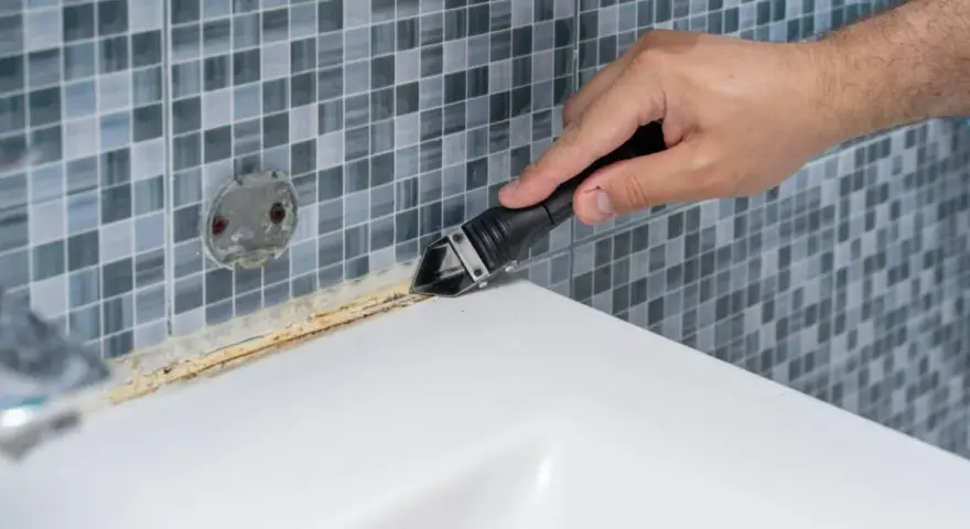 Mano usando una espátula para quitar silicona vieja y sucia de la bañera.