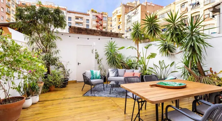 Un patio exterior pequeño transformado con una reforma: suelo de madera, muebles de exterior, plantas exuberantes y luces decorativas crean un oasis urbano.