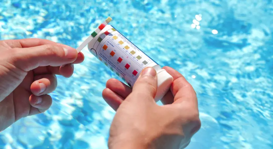 Manos sostienen una tira reactiva para medir el pH de la piscina. El agua azul y brillante de la piscina se ve al fondo.