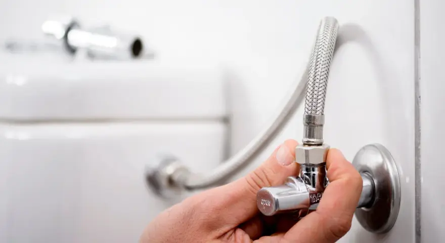 Mano ajustando uno de los tipos de llaves de paso de agua, conectada a una manguera flexible y al tanque de un inodoro.