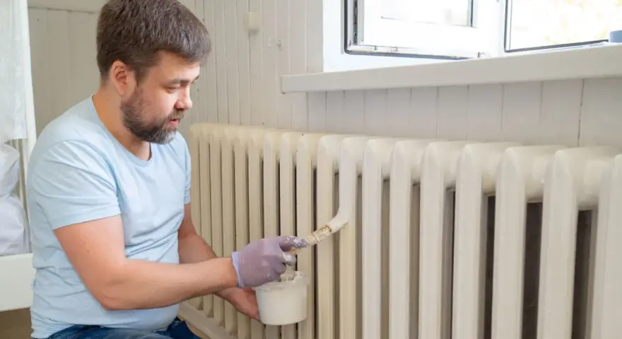 Hombre con barba pintando radiadores blancos con brocha y pintura.
