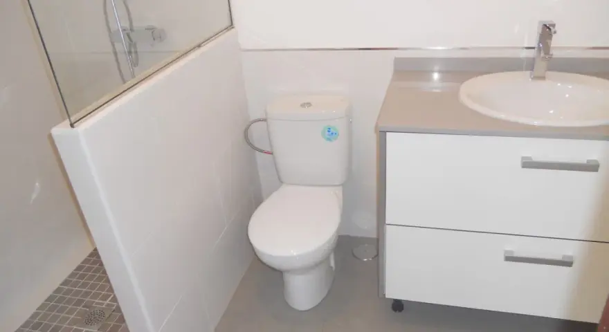 Baño moderno con ducha separada por muro de azulejos blancos y cristal. Inodoro y lavabo sobre mueble blanco.