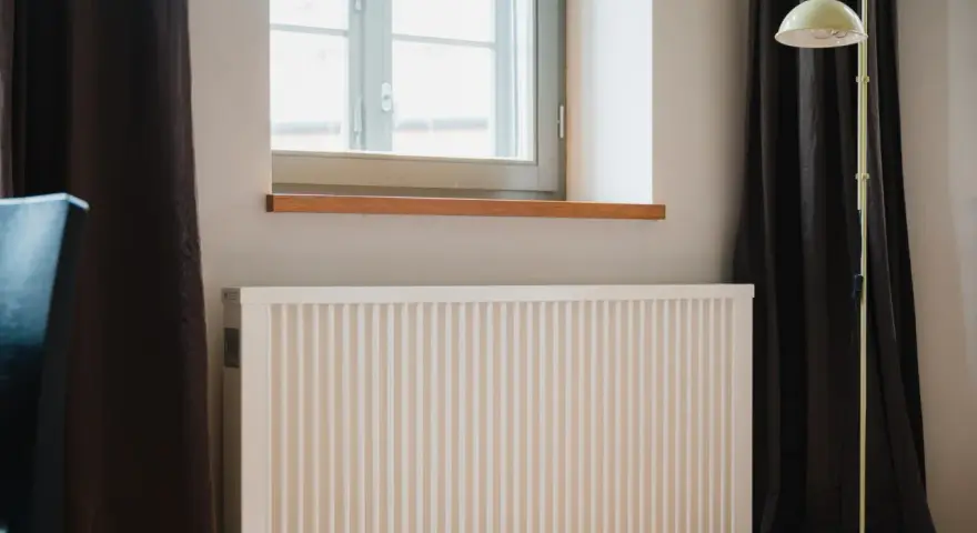 Radiador blanco bajo ventana, con cortinas oscuras y lámpara amarilla. El **calor azul** del invierno se combate con este sistema.