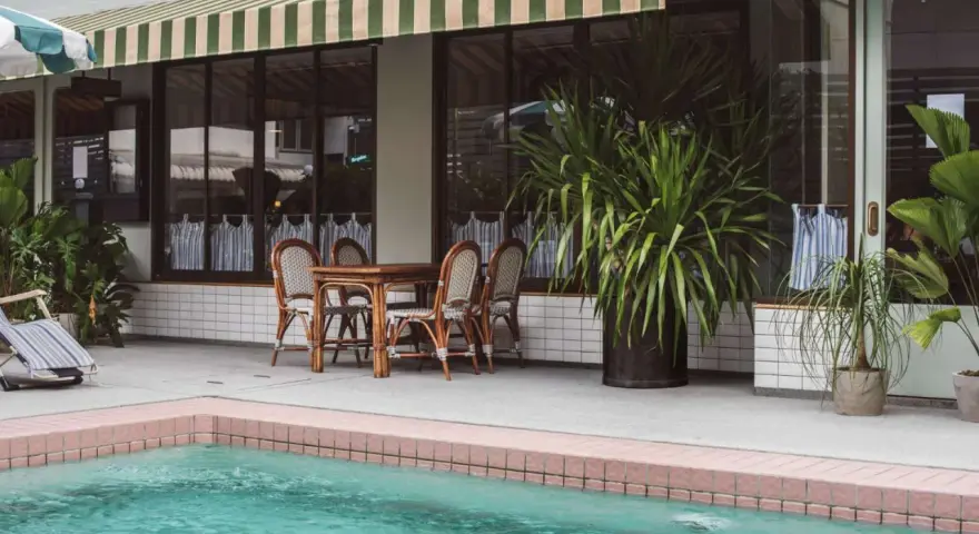 Terraza con piscina y toldo a rayas verdes y blancas. Mesa de madera con sillas de mimbre listas para instalar toldo.