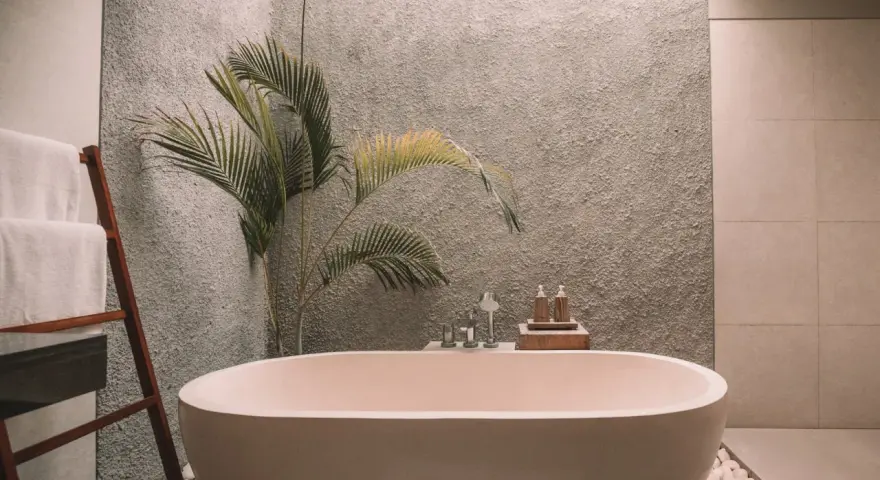 Un baño moderno con una bañera ovalada, toallas blancas en una escalera de madera y una planta de hojas verdes. La pared tiene textura y el suelo está cubierto de guijarros. ¡Listo para pintar la bañera y renovar el espacio!