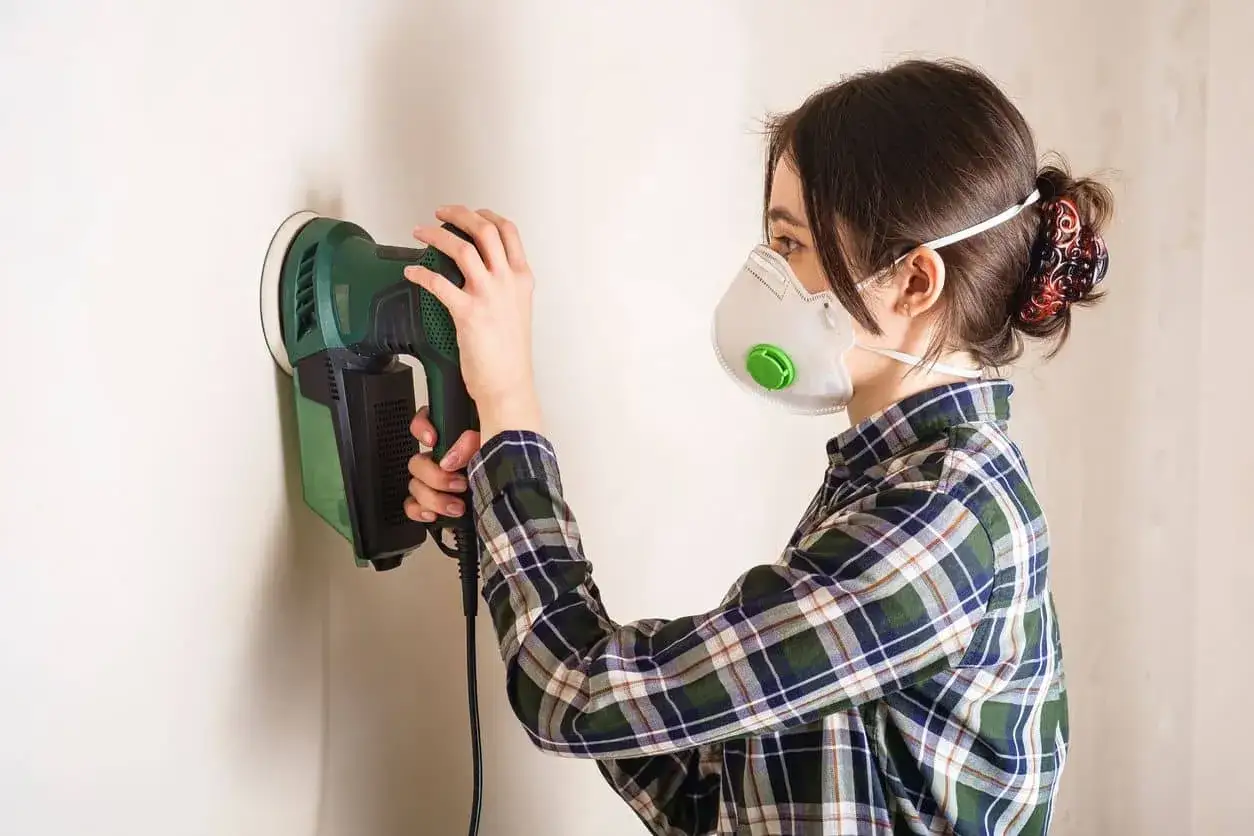 Mujer con mascarilla lijando paredes con una lijadora el&eacute;ctrica.