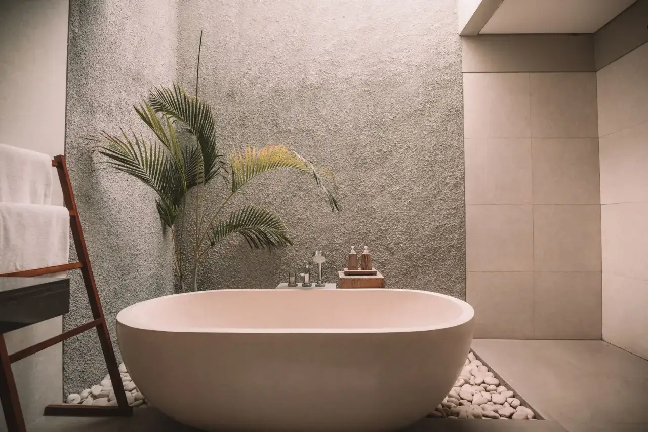 Bañera independiente en un spa moderno, rodeada de piedras blancas y vegetación. Toallas blancas en un tendedero de madera.