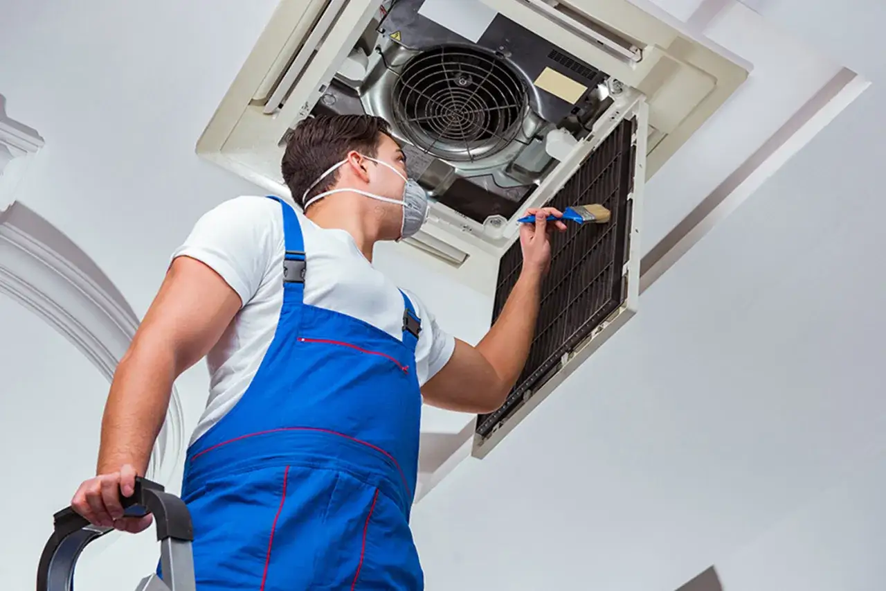 Técnico con mascarilla realiza mantenimiento de aire acondicionado, limpiando el filtro con un cepillo.