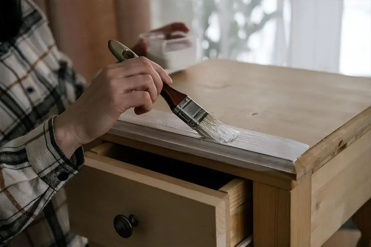 Mano con pincel pintando de blanco la madera de un mueble.
