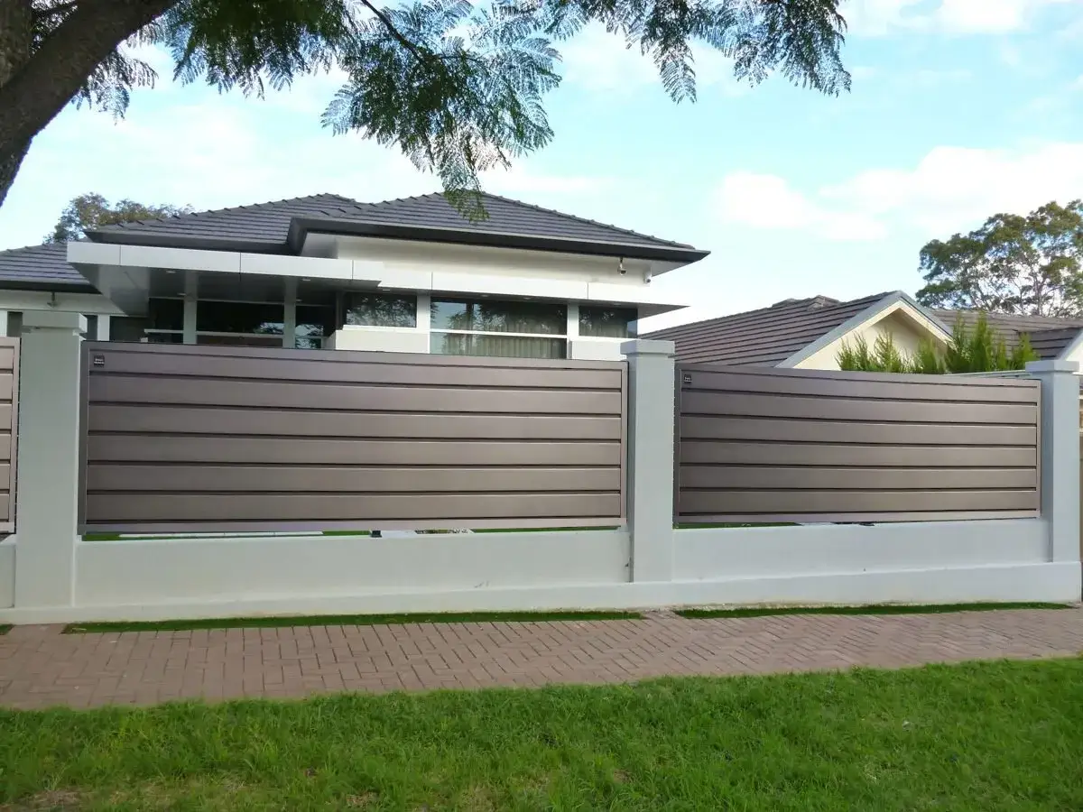 Moderna verja jardin de listones horizontales grises, con pilares blancos y base de hormig&oacute;n. Casas modernas al fondo.