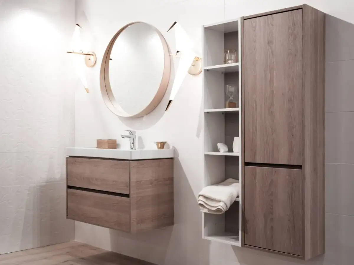 Modern bathroom with a white sink and wooden vanity. A round mirror hangs above, and a tall cabinet with shelves is to the right.