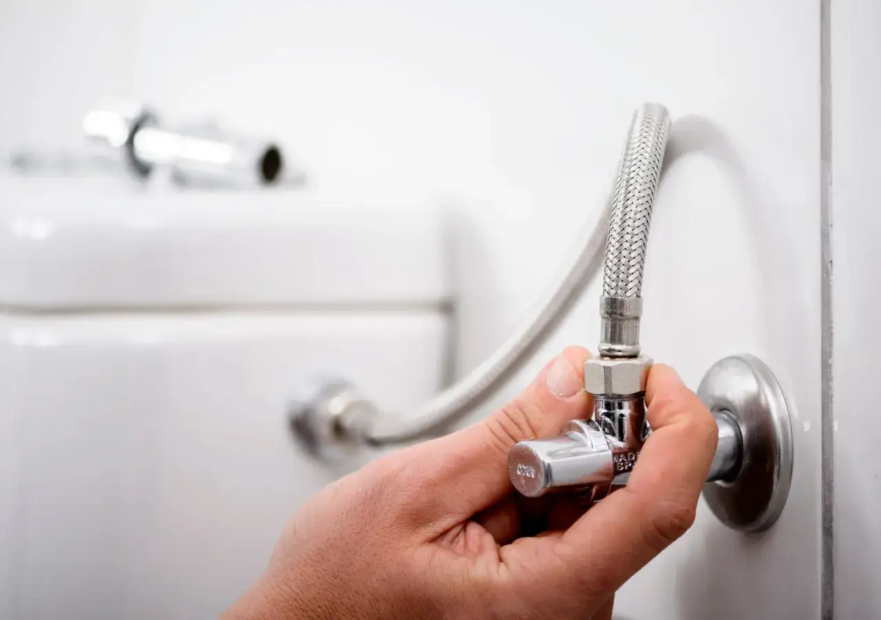 Mano ajustando uno de los tipos de llaves de paso de agua, conectada a una manguera flexible y a la pared.