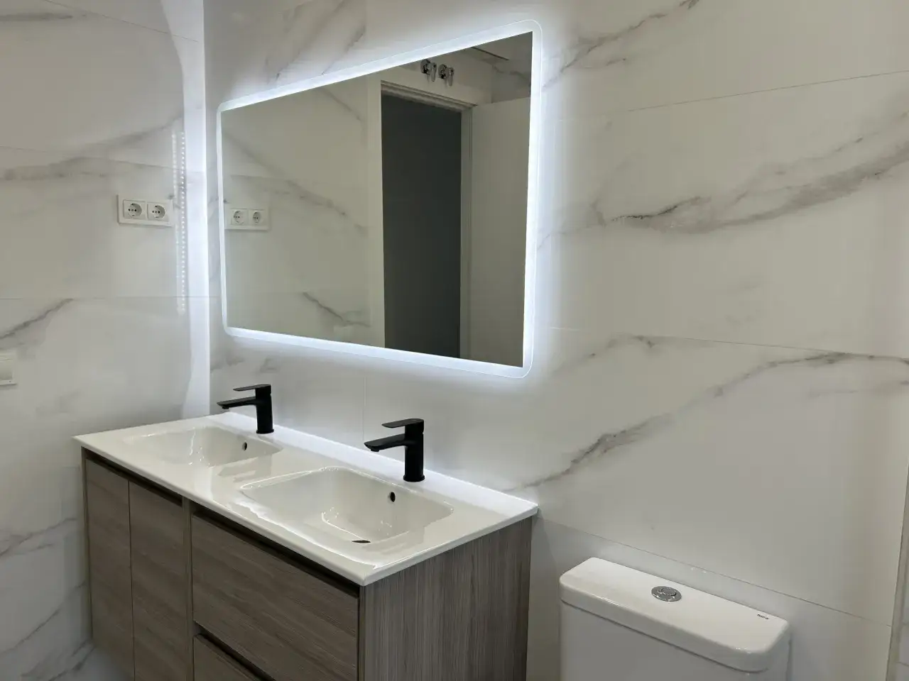 Moderno ba&ntilde;o con doble lavabo y grifer&iacute;a negra. Las paredes lucen un elegante revestimiento imitando m&aacute;rmol, una soluci&oacute;n ideal para revestimiento paredes ba&ntilde;o sin obras.