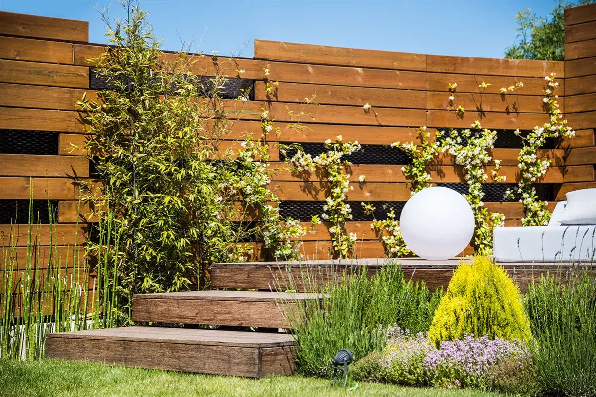 Un moderno jard&iacute;n con una verja de madera, escalones de madera, plantas verdes y una esfera luminosa.