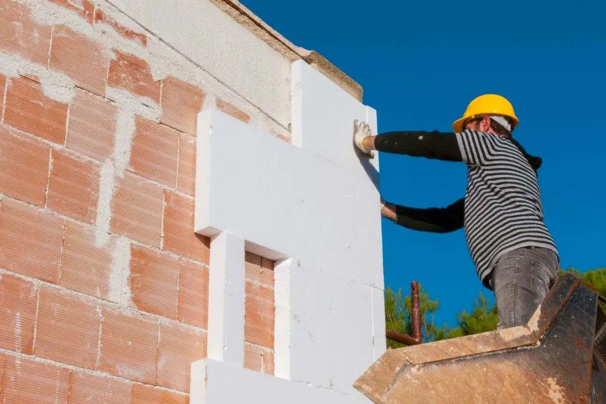 Obrero con casco amarillo instala paneles de poliestireno para aislar fachadas.