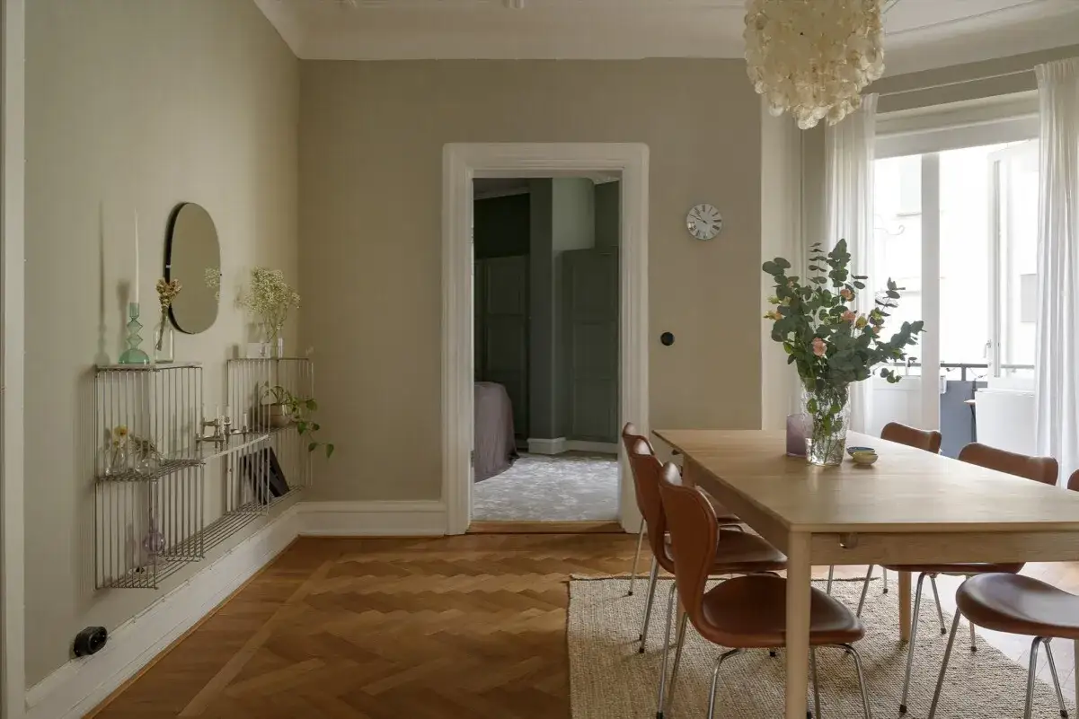 Comedor con mesa de madera, sillas marrones y pared color crema. Un jarrón con flores decora la mesa.