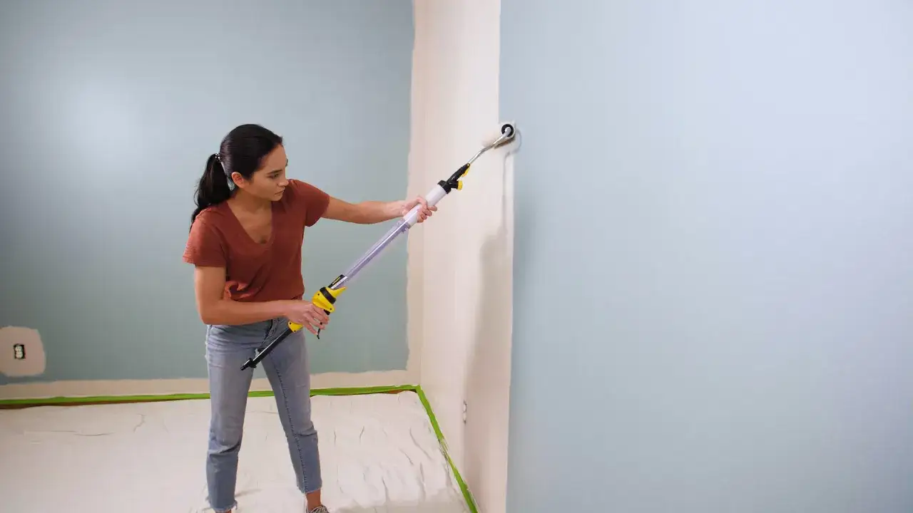 Mujer pinta pared con rodillo extensible. La pintura al agua cubre la superficie con un tono azul claro.