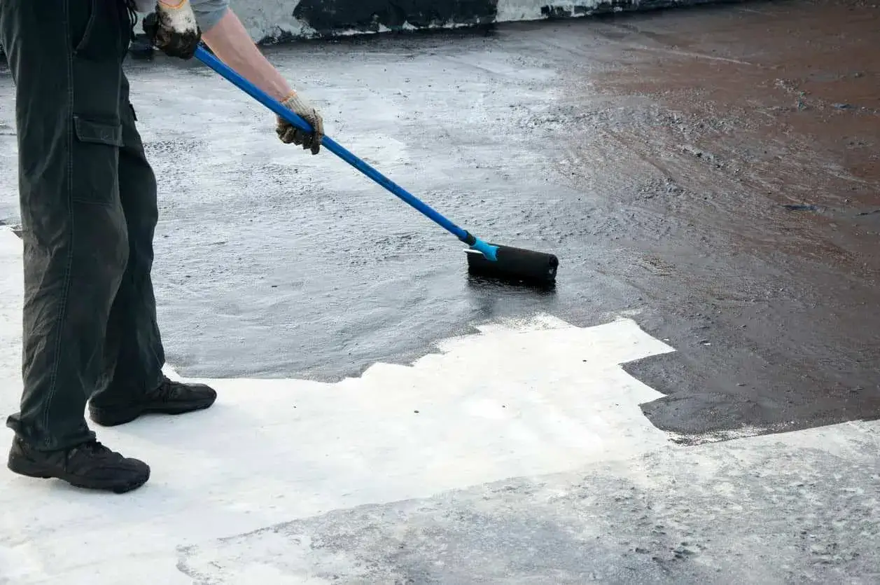 Persona aplica pintura oscura con rodillo sobre suelo de cemento sin pulir, cubriendo una zona blanca.