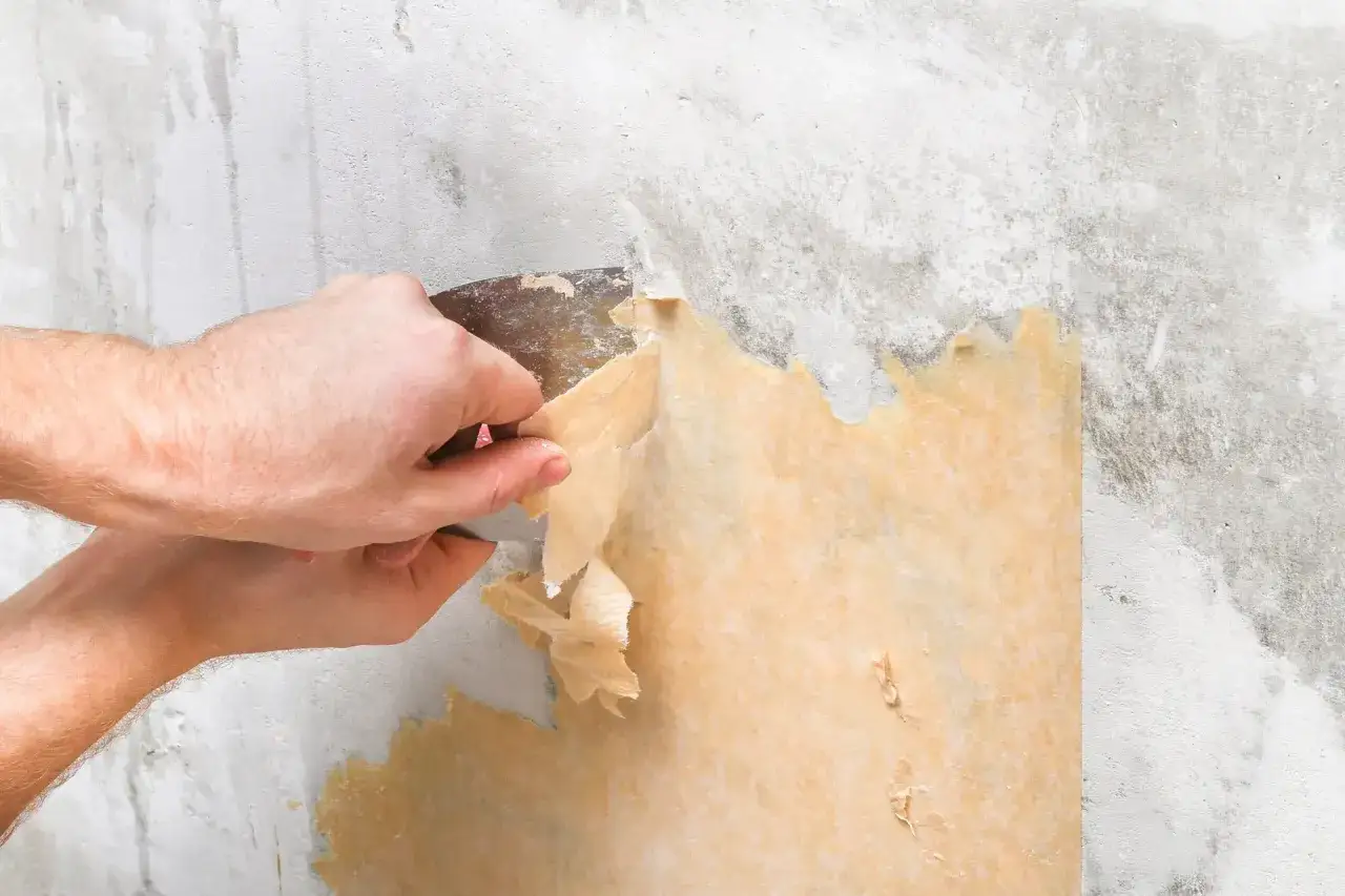 Manos usando una esp&aacute;tula para quitar papel pintado viejo de una pared gris.