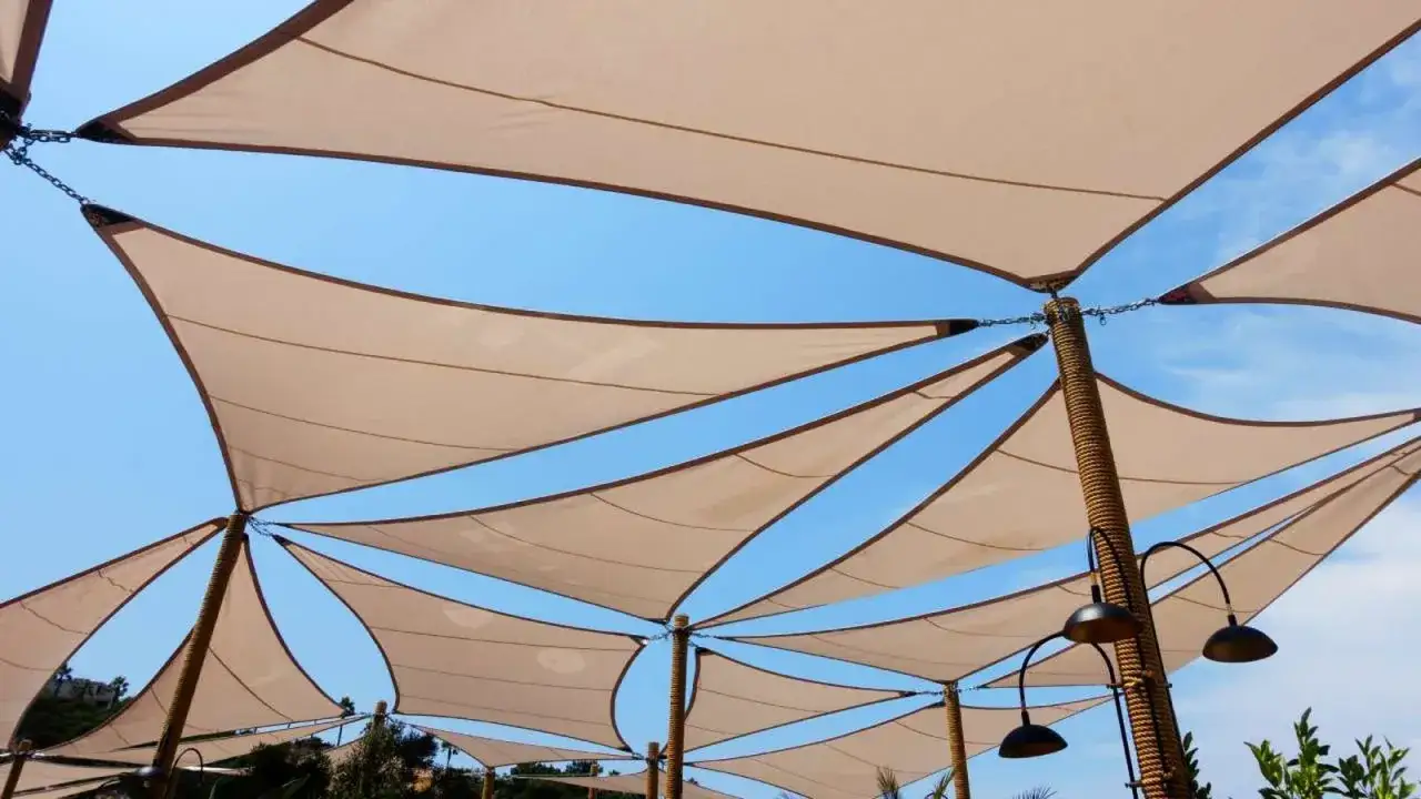 Instalar toldo para crear sombra en un d&iacute;a soleado. Las velas de tela beige se extienden contra el cielo azul, sujetas a postes cubiertos de cuerda.