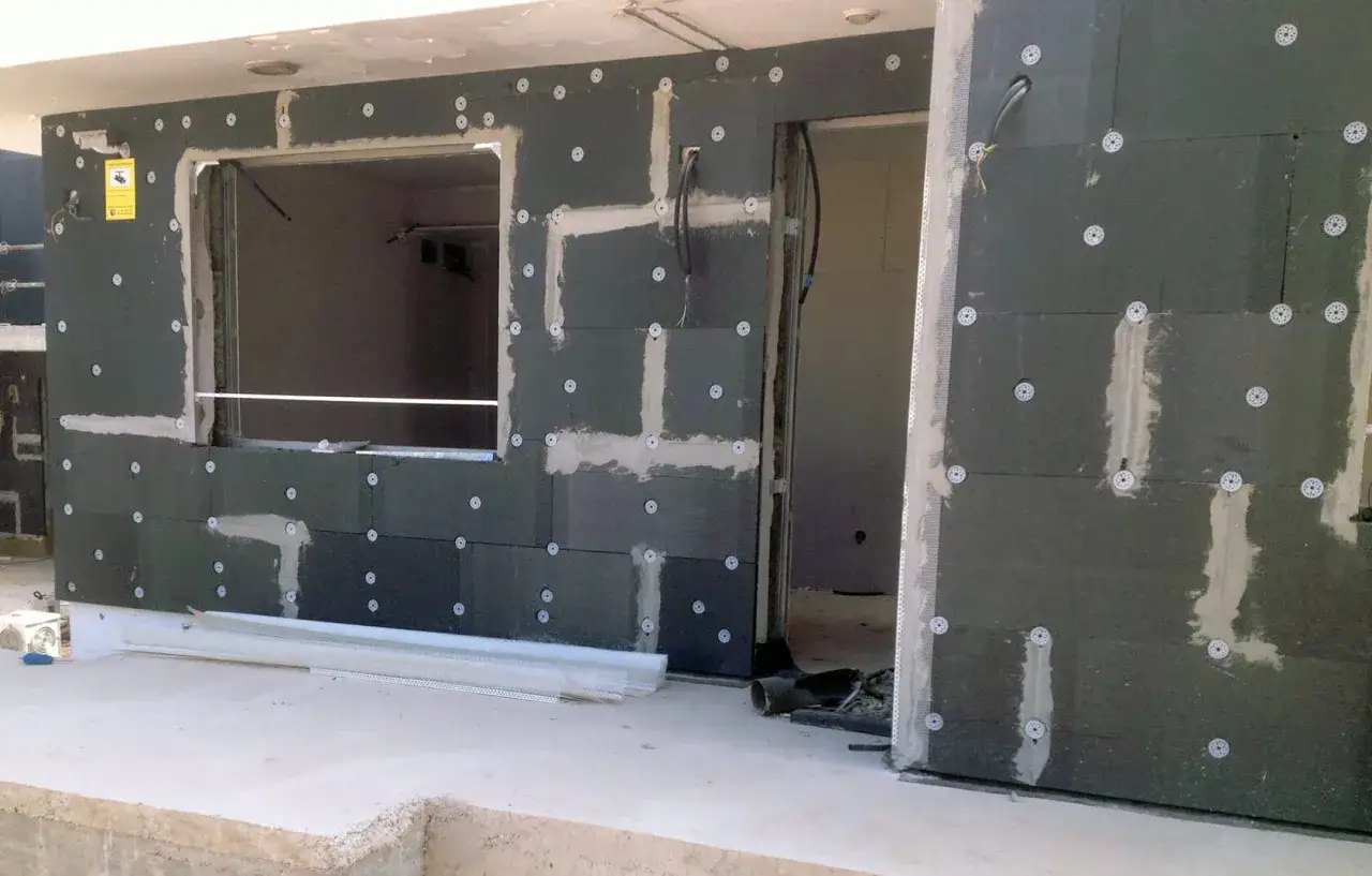 Paneles de aislante t&eacute;rmico exterior fijados a una pared con mortero y discos. Se aprecian un hueco para ventana y una entrada.